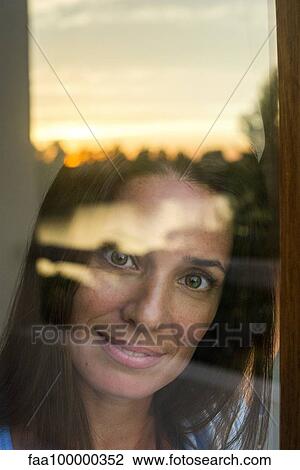 Stock Image - Woman looking through window at camera, portrait. Fotosearch