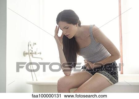 Stock Image - Woman sitting on edge of bathtub holding head and stomach. Fotosearch