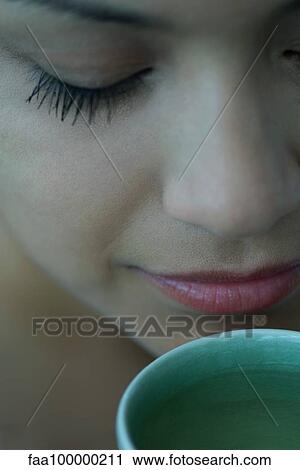 Stock Image - Woman smelling cup of tea with eyes closed, close-up. Fotosearch