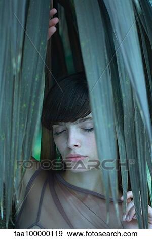 Stock Photo - Woman standing among palm leaves, eyes closed. Fotosearch