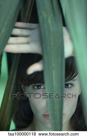 Stock Photo - Woman standing behind palm leaves, looking at camera. Fotosearch