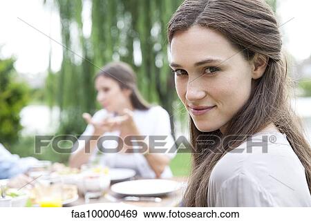 Young woman sitting at table outdoors, smiling over shoulder, portrait View Large Photo Image Stock Photo - Young woman sitting at table outdoors, smiling over shoulder, portrait. Fotosearch