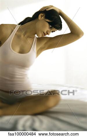 Picture - Young woman sitting on bed stretching, hand on head. Fotosearch