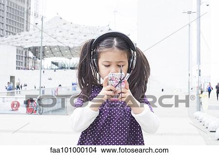 Picture - Girl looking at smartphone and listening to headphones outdoors. Fotosearch