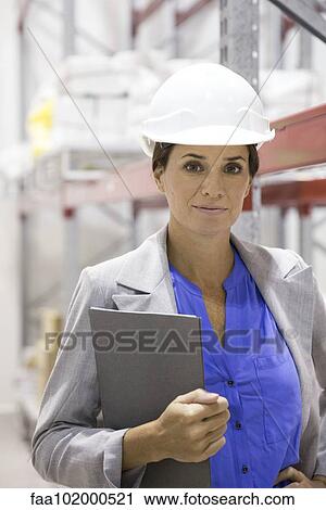 Stock Image - "Businesswoman with hard hat in warehouse, portrait". Fotosearch