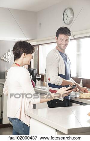 Stock Photo - Couple looking at smartphone together while preparing food in kitchen. Fotosearch