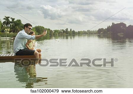 Stock Photo - Father and daughter sitting on dock taking selfie. Fotosearch