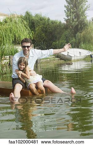 Stock Image - "Father and young daughter sitting on dock, portrait". Fotosearch