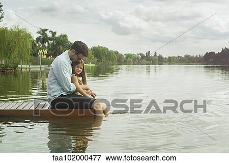 Stock Photo - Father and young daughter sitting on dock with legs dangling in lake. Fotosearch