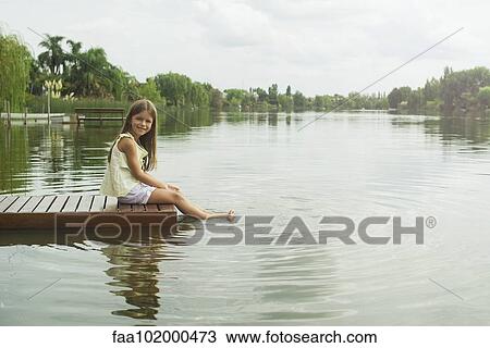 Stock Image - "Girl sitting on dock with feet dangling in lake, portrait". Fotosearch