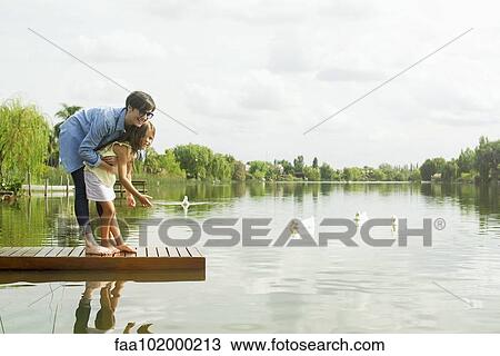 Stock Image - Mother and young daughter feeding ducks at lake. Fotosearch