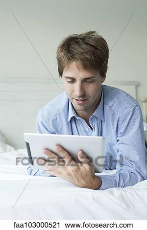 Stock Image - Man lying in bed using digital tablet. Fotosearch
