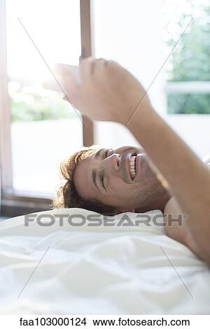 Picture - Man lying in bed using multimedia smartphone for instant messaging. Fotosearch