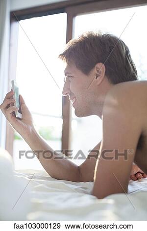 Man lying in bed using multimedia smartphone to video chat View Large Photo Image Stock Photo - Man lying in bed using multimedia smartphone to video chat. Fotosearch