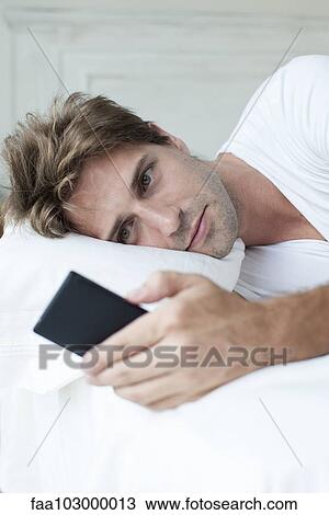 Stock Image - Man lying in bed using smartphone. Fotosearch