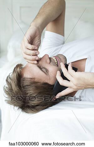 Stock Image - Man talking on cell phone on bed. Fotosearch