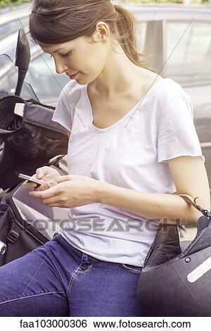Stock Photograph - "Woman leaning against motorcycle, text messaging with cell phone". Fotosearch