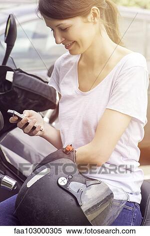 Stock Photography - "Woman sitting on motorcycle, smiling at smartphone". Fotosearch