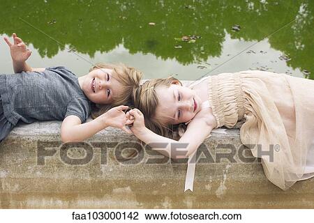 Stock Image - "Young sisters lying down on beside pond, holding hands". Fotosearch