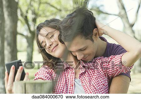 Stock Image - Couple embracing outdoors, posing for a selfie. Fotosearch