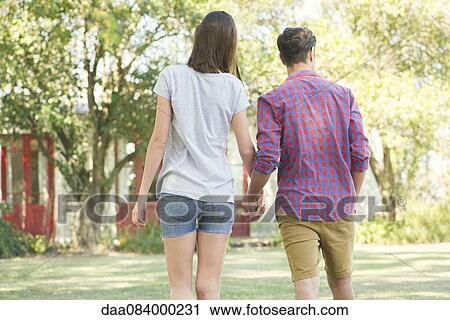 Stock Image - Couple walking and holding hands outdoors, rear view. Fotosearch