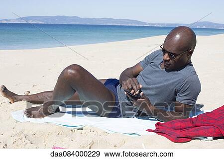 Stock Photo - Man lying on beach, using smartphone. Fotosearch