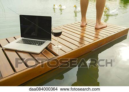 Picture - Man standing beside laptop computer and wine glass on lake pier. Fotosearch