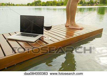 Stock Image - Man standing beside laptop computer and glass of wine on pier. Fotosearch