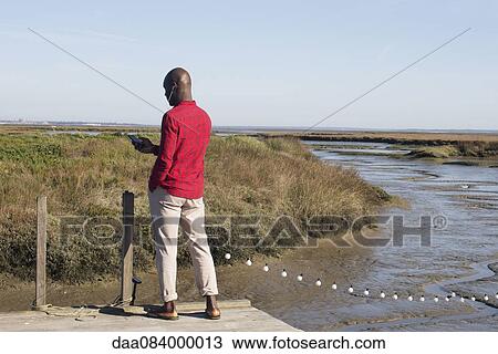 Stock Image - Man standing on dock, listening to music with earphones. Fotosearch