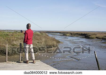 Stock Image - Man standing on dock, looking at multimedia smartphone. Fotosearch