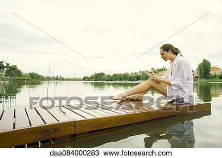 Stock Image - Woman sitting on lake dock using digital tablet. Fotosearch