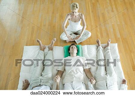 Picture - Alternative therapy session, three adults lying side by side, holding hands while therapist puts hands over man's head. Fotosearch