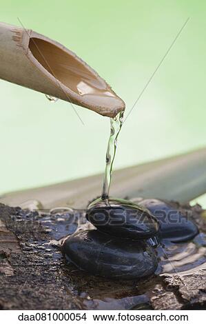Picture - Bamboo spout pouring water over stack of black pebbles. Fotosearch