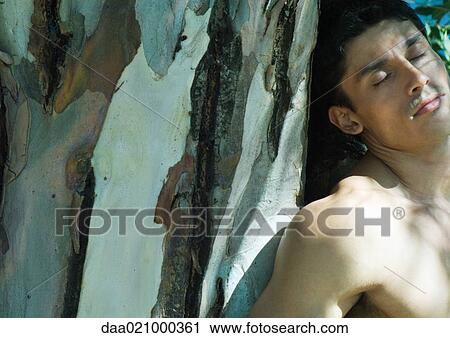 Stock Image - Barechested man leaning against tree trunk. Fotosearch