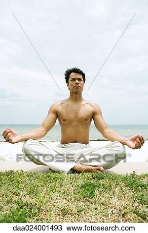 Stock Image - Barechested man sitting in lotus position, ocean in background, full length. Fotosearch
