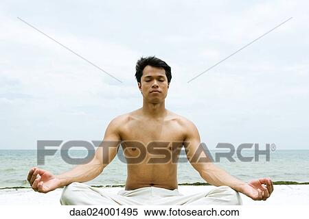 Stock Photography - Barechested man sitting in lotus position, ocean in background. Fotosearch