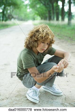 Stock Photo - Boy crouching in middle of dirt road, looking at pebbles. Fotosearch