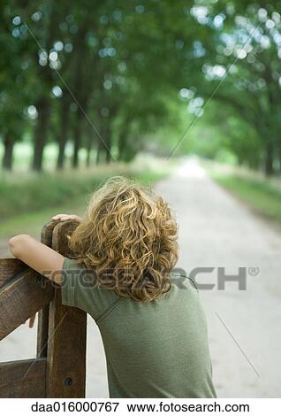 Stock Photo - Boy leaning on gate, facing dirt road, rear view. Fotosearch