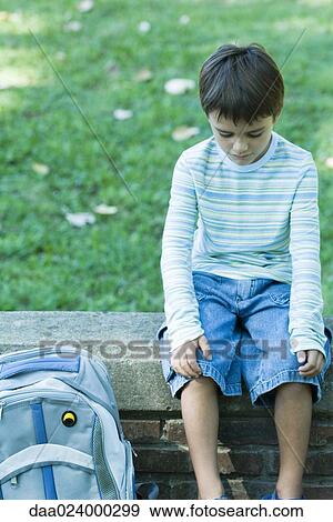 Stock Photo - Boy sitting on low wall, backpack by side, looking down. Fotosearch