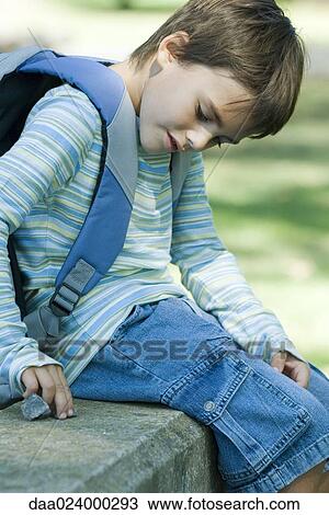 Boy sitting on low wall, wearing backpack, playing with pieces of gravel View Large Photo Image Stock Image - Boy sitting on low wall, wearing backpack, playing with pieces of gravel. Fotosearch