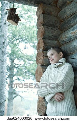 Stock Photograph - Boy wearing wool sweater, arms folded, leaning against log cabin. Fotosearch