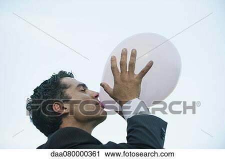 Stock Image - Businessman blowing up balloon, side view. Fotosearch
