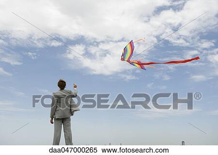 Stock Photography - Businessman flying kite in field, rear view. Fotosearch