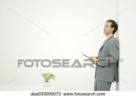 Stock Image - Businessman leaning against wall and holding document, looking away. Fotosearch