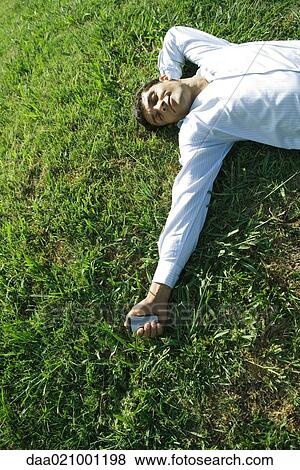 Stock Photo - Businessman lying on grass, holding cell phone, eyes closed, smiling. Fotosearch