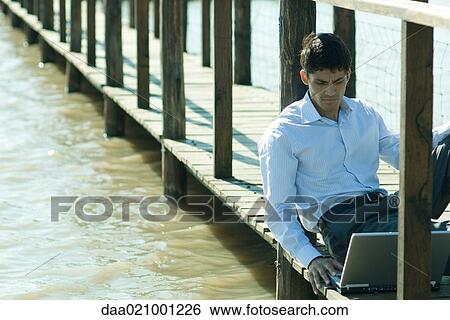 Businessman on wooden footbridge over lake, using laptop View Large Photo Image Stock Photograph - Businessman on wooden footbridge over lake, using laptop. Fotosearch