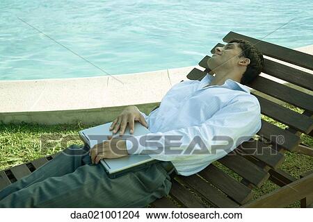 Picture - Businessman reclining in lounge chair near pool, holding laptop on lap. Fotosearch