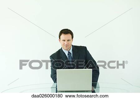 Stock Image - Businessman sitting at desk, using laptop computer, smiling at camera. Fotosearch