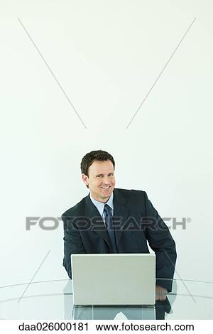 Businessman sitting at desk, using laptop computer, smiling at camera View Large Photo Image Stock Image - Businessman sitting at desk, using laptop computer, smiling at camera. Fotosearch