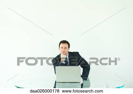 Businessman sitting at desk with laptop computer, hand under chin, looking up View Large Photo Image Stock Photo - Businessman sitting at desk with laptop computer, hand under chin, looking up. Fotosearch